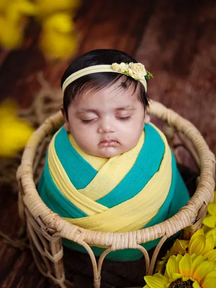 A baby wrapped in a yellow and teal swaddle rests in a simple basket, surrounded by sunflowers.