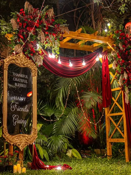 A cozy and stylish Friendsgiving dinner in a garden. The decor featured a custom mirror sign, a wooden arch with rich red drapery, and plenty of warm lighting.