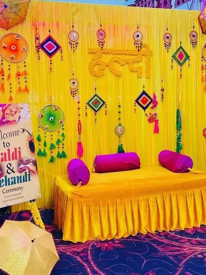 A vibrant Haldi and Mehendi seating area with a yellow velvet-covered diwan, a colorful backdrop, and a welcome sign for guests.