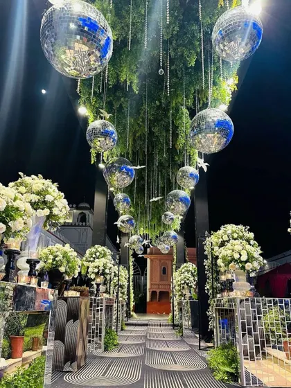An enchanting walkway for a reception, with a ceiling of hanging greenery, crystal strings, and disco balls that reflect light beautifully. The path is lined with mirrored pedestals and white flowers.