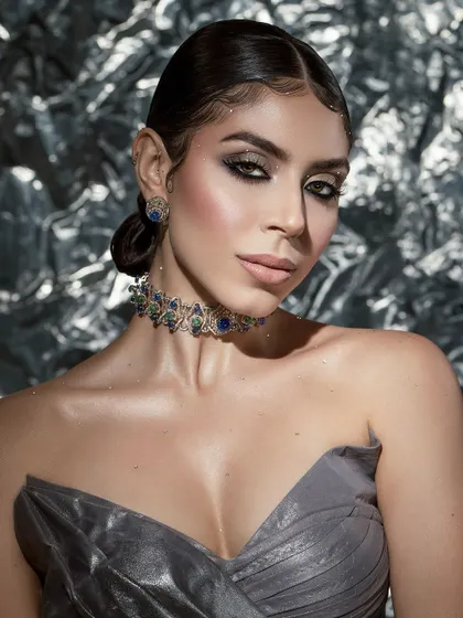 A high-glam beauty portrait against a metallic background. The focus is on the sculpted features, smoky eyes, and the exquisite sapphire choker.