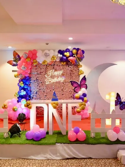 A first birthday party with a butterfly and moon theme. The setup includes a white picket fence, a large moon prop, a sequin backdrop, and "ONE" table.