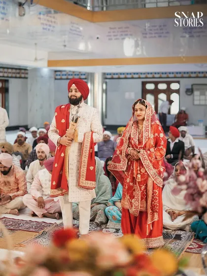 The couple stands together during the Anand Karaj ceremony. This full-length shot captures the elegance of their coordinated red and white wedding outfits.