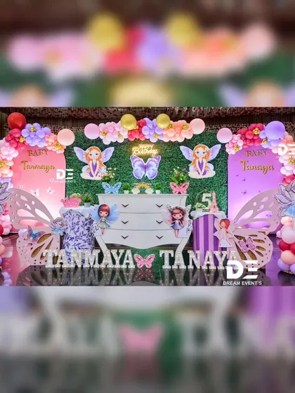 A magical fairy-themed party for twins Tanmaya and Tanay. The setup includes a green hedge backdrop, a white dresser as a centerpiece, and enchanting fairy and butterfly cutouts, all tied together with pink and purple balloons.