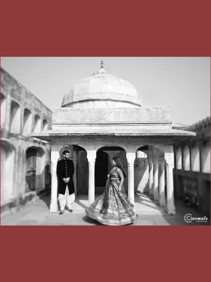 A black and white photograph taken in a palace courtyard. The woman's twirling lehenga adds a sense of movement and joy, contrasting beautifully with the static, grand architecture around them.