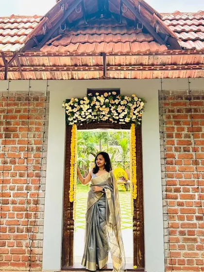A full-length shot of Maneesha in her elegant grey saree, standing in a beautifully decorated doorway. A perfect blend of tradition and contemporary style.