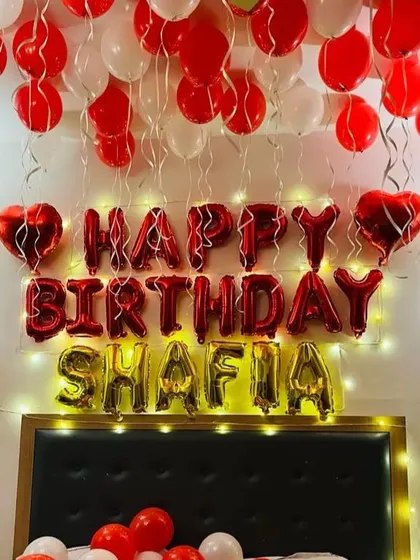 A personalized birthday decoration over a bed. The setup includes red and white balloons on the ceiling, 'Happy Birthday' and name foil letters, and fairy lights.