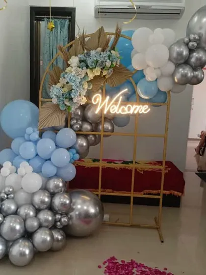 A stylish welcome decor piece. The gold grid frame is decorated with a blue, white, and silver balloon arrangement and a "Welcome" neon sign.