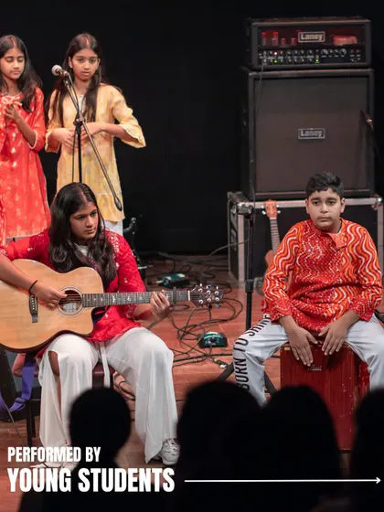 A group of students performs at 'Holiday in Harmony', with one playing guitar, another on cajon, and a choir singing. Our musicals showcase a wide array of instruments and talents.