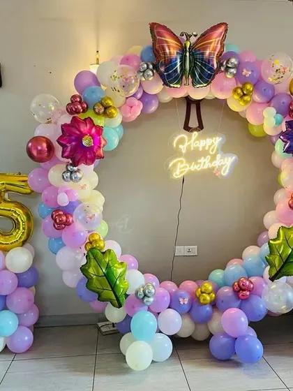 A colorful 5th birthday party with a circular balloon arch, butterfly and flower foils, and a 'Happy Birthday' neon sign.