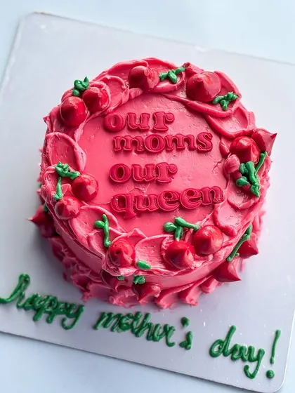 A special cake for Mother's Day with the message "Our Moms Our Queen." The vibrant pink buttercream is decorated with piped cherries for a fun and festive look.