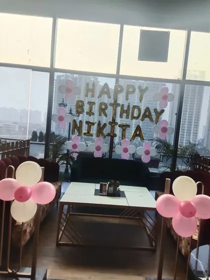 A sweet and simple birthday decoration for 'Nikita', using pink and white flower-shaped balloon arrangements and gold foil letters against a large window.