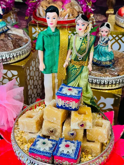 A sweet platter featuring a traditionally dressed couple doll. The tray is filled with jaggery blocks and decorated with small, ornate boxes, perfect for a wedding ceremony.
