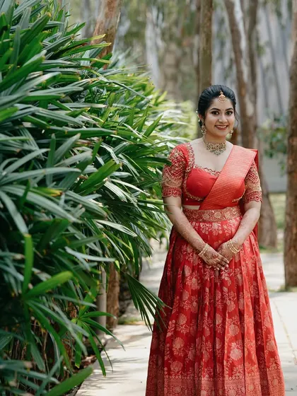 A full-length shot of the bride in her red brocade half-saree. The makeup is elegant and not overpowering, letting her natural beauty and outfit be the focus.