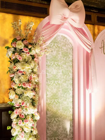 A beautiful floral arch in shades of pink and white, accented with a large pink bow, part of a 'Bow'teaful' party theme.