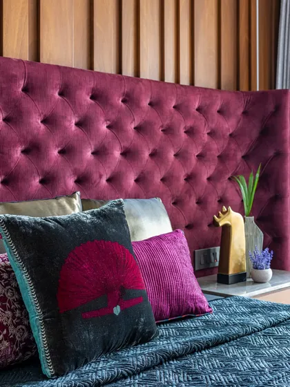 A close-up of the plush, tufted burgundy headboard in the Filigree House. The rich texture and color, paired with curated cushions, make this bed the undeniable centerpiece of the room.