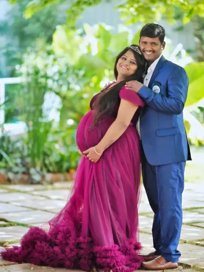 A happy couple posing together during an outdoor garden photoshoot. The partner's blue suit coordinates beautifully with the deep magenta of the maternity gown.