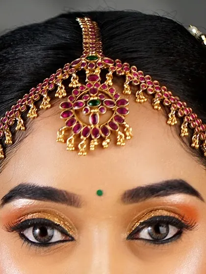 A macro shot of bridal eye makeup. The focus is on the precise application of the golden glitter eyeshadow and the traditional red and green matha patti.