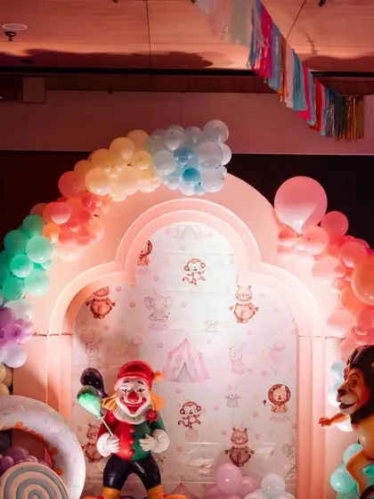 A fun photo backdrop with a rainbow balloon arch and clown and lion props. The wallpaper features cute, subtle circus animal illustrations.