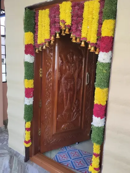 A door decoration featuring a 'bell design' with flower garlands and brass bells. This is a popular choice for Gruha Pravesha ceremonies.