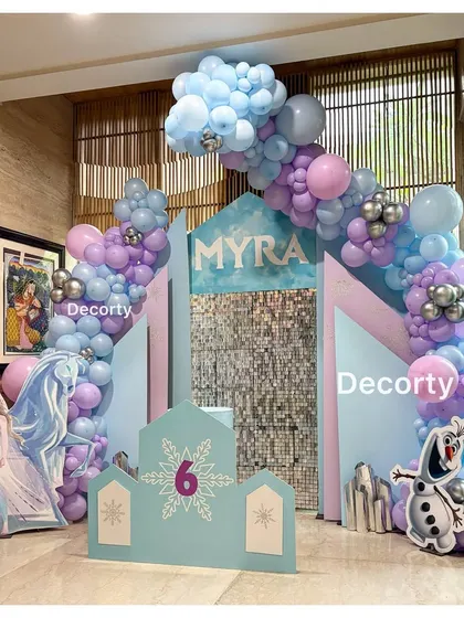 A magical 'Frozen' themed birthday backdrop for Myra's 6th birthday. The setup includes an icy castle, cutouts of Elsa and Olaf, and a shimmering sequin wall, all framed by a blue and purple balloon arch.
