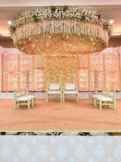 A full view of a Vidhi Mandap designed in soft pastel tones. The stage features a ceiling completely covered in hanging flowers and a backdrop of shimmering gold sequins, creating a space that feels both traditional and dreamlike.
