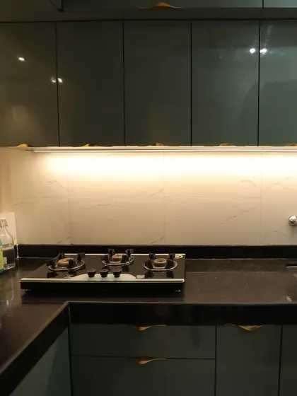 A modular kitchen with high-gloss teal cabinets and modern G-profile handles. The under-cabinet lighting highlights the white marble-patterned backsplash.