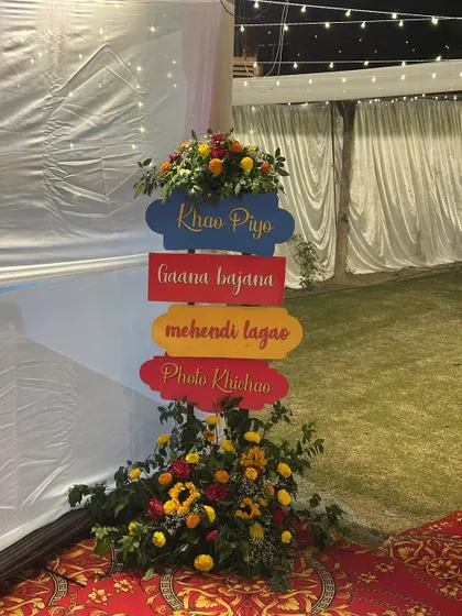 A colorful and floral directional signpost for a Haldi event. The signs, adorned with flowers, guide guests to different activities like 'Khao Piyo' and 'Mehendi Lagao'.