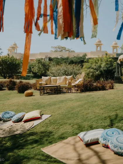 A relaxed, picnic-style seating area on the lawn. Mats and printed cushions invite guests to unwind under a tree adorned with flowing ribbons, with the grand Udaivilas palace in the background.