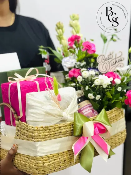 A large, impressive gift basket being held, ready for delivery. It's overflowing with fresh tuberoses and pink flowers, beautifully wrapped gifts, and a "Best Wishes" sign, perfect for any major celebration.