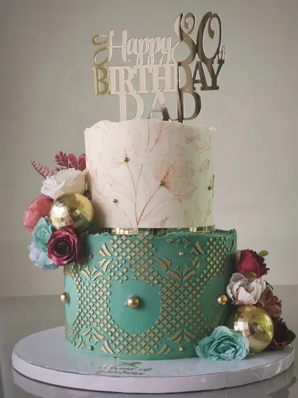A distinguished two-tiered cake for an 80th birthday. The design includes a hand-painted floral top tier and a bottom tier with a green and gold geometric pattern, creating a look of timeless elegance.