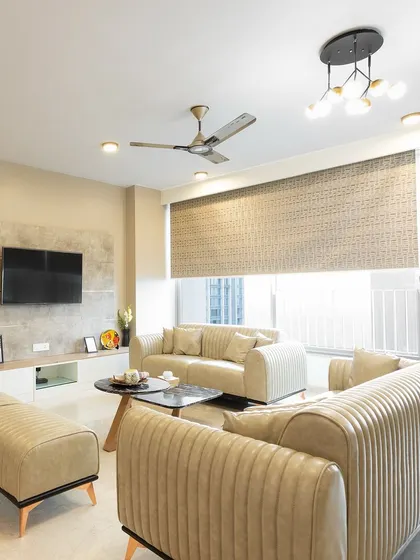 A wide view of the comfortable living room with beige sofas, a simple entertainment unit, and large windows with blinds for light control.