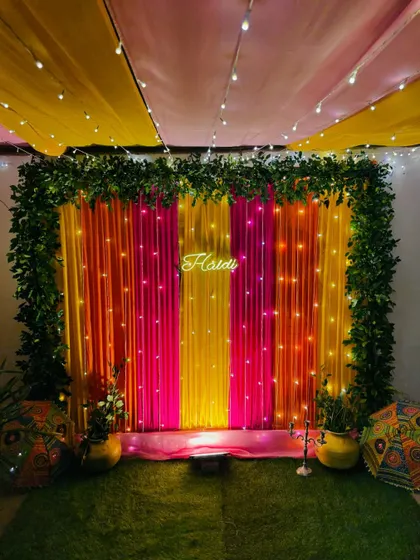 A colorful Haldi backdrop for a home event. The decor features a frame of green leaves around pink and yellow drapes, with a 'Haldi' neon sign as the centerpiece.