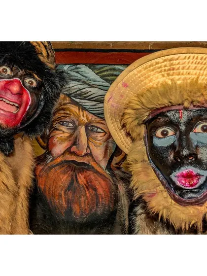 A striking street portrait featuring two performers in expressive masks and makeup. The image plays with layers of identity, contrasting the live faces with the painted face in the background, creating a compelling visual narrative.