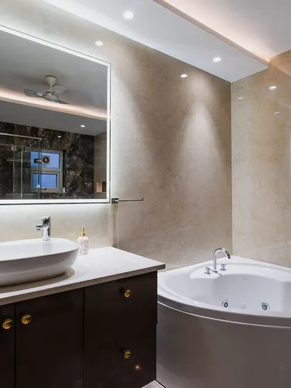 This bathroom is all about indulgence. It features a corner jacuzzi tub, a large backlit mirror, and a spacious vanity. The beige and brown marble tiles create a warm and inviting atmosphere.