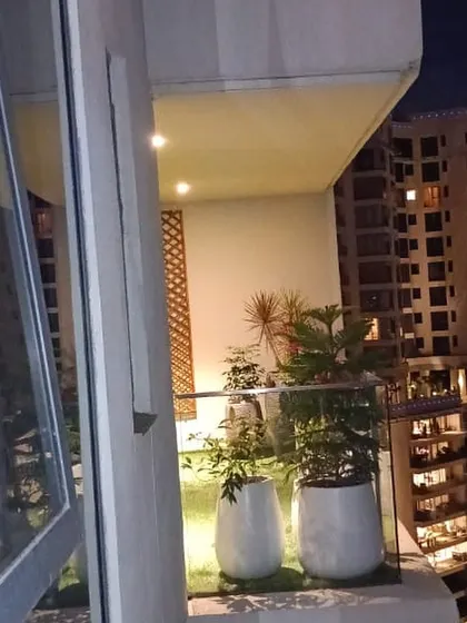 A view from inside looking out onto a high-rise balcony at night. The artificial grass and plants in white pots are softly lit, creating an inviting green space against the city lights.