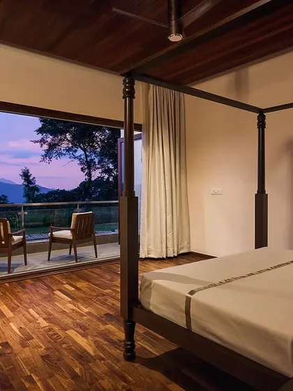 A serene bedroom featuring a classic four-poster bed and a private sit-out. The space is designed to be a peaceful escape, with the evening sky and distant mountains providing a tranquil backdrop.