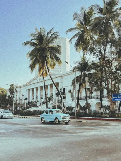 A classic light blue Fiat driving past the Asiatic Library, with palm trees framing the shot. This feels like a scene from an old movie.