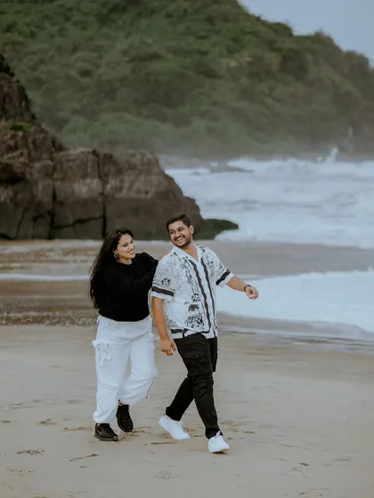 A candid shot of a couple walking and laughing on a sandy beach, with dramatic cliffs and waves in the background.
