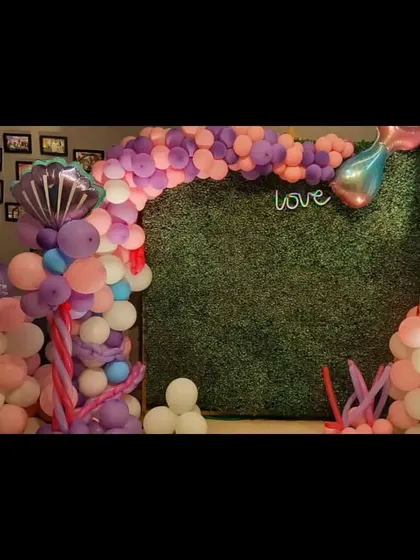 A magical mermaid theme birthday decoration. The setup features a green backdrop with a pink and purple balloon arch and mermaid tail accents.