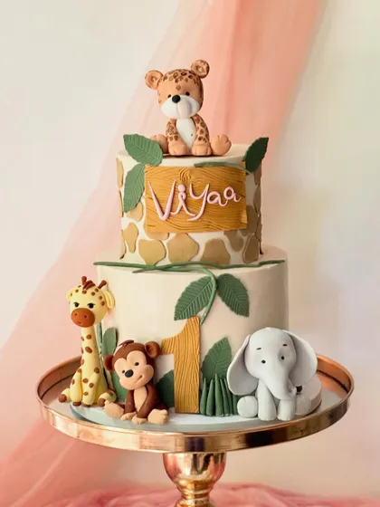 A two-tier jungle-themed cake with a giraffe print pattern on the top tier. The cake is adorned with a leopard, giraffe, monkey, and elephant for a wild first birthday.