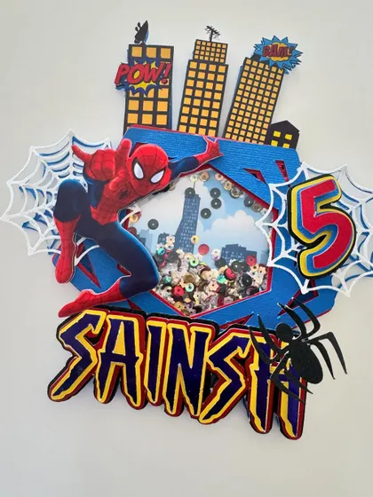 A full view of the handmade Spiderman shaker topper. The layered design includes 3D buildings, spider webs, and a personalized name in a comic-book font.