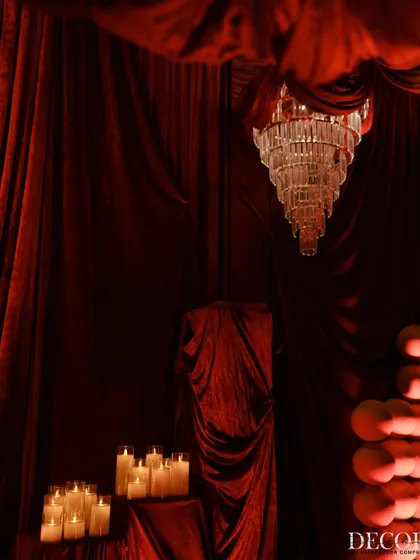 A composition of light and shadow, featuring a grand chandelier, pillar candles, and textured red spheres against a backdrop of velvet drapes.