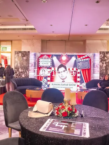 The main event space for a Bollywood-themed 90th birthday party. The setup includes a custom stage designed to look like a film set, cocktail tables with sequined cloths, and a large screen for presentations.