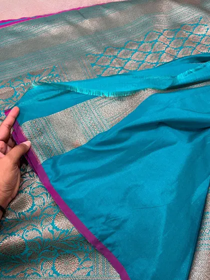 The plain teal blouse piece with a simple magenta border, designed to complement the heavily woven saree.