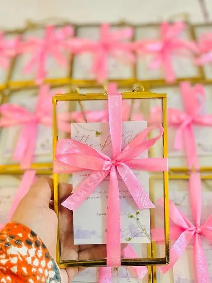 These elegant wedding invites for Apoorva and Aditya feature a brass photo frame holding the invitation card, tied with a pink ribbon.