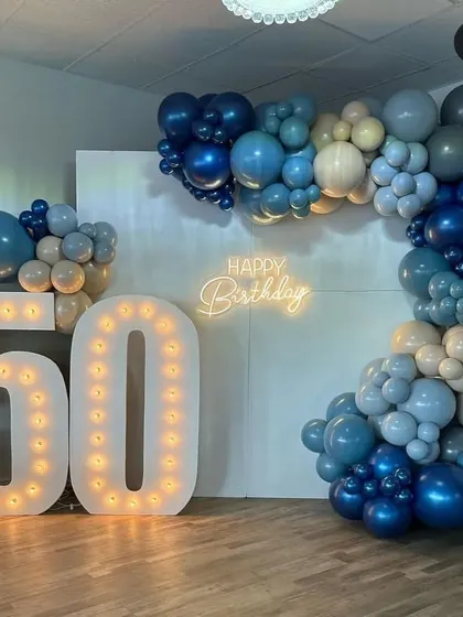 Milestone birthdays deserve a grand statement. This 50th birthday decoration features large, illuminated marquee numbers, a custom 'Happy Birthday' neon sign, and a stylish balloon garland in a cool palette of blues, greys, and beige.