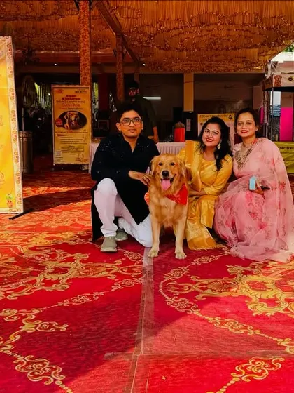 A wide shot of Roger and his family at the Durga Puja event, showing the festive setup with stalls and banners in the background.