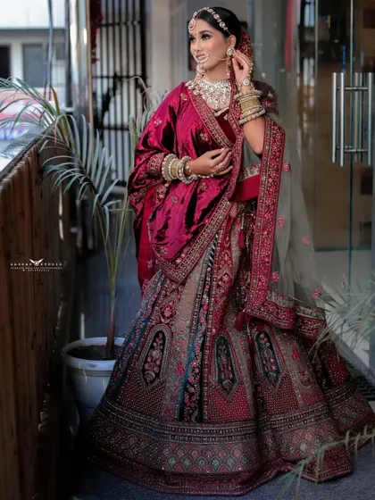 A candid-style shot of the bride smiling, captured outdoors. This photo highlights the luxurious texture of her maroon velvet lehenga and her natural, happy expression.
