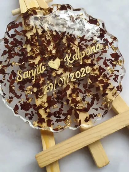 A close-up of a single coaster made from preserved flower petals and gold leaf. Personalized with the couple's names, it serves as a small, beautiful reminder of their special day.